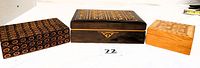 Photo showing three small wooden boxes: dark wood carved box with foreign writing, a box with fire decoration, and a light-colored Marquetry trinket box with quilt pattern.