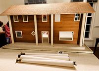 Front view of the vintage wooden dollhouse showing shingle roof, white columns, window openings, and four detachable legs laid in front.