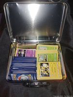 Open metal collector's tin filled with stacks of Pokemon cards with blue, yellow, green, and purple backs and fronts visible, showing Electrike and other characters.