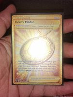 Full front view of the Hero's Medal Pokémon card showing gold foil holographic design and text details