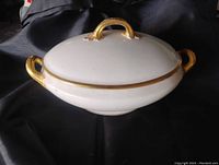 White oval porcelain serving dish with lid and gold handles and rim decoration on black background