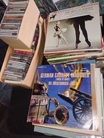 Photo showing stacks of sealed CDs in plastic cases stored in cardboard boxes and two prominent 12-inch vinyl records on top of each stack.