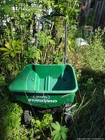Front view of green Scotts Standard seed spreader with the bucket, handlebar, and wheels visible; placed outside in garden area.