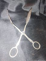 Silver plated lifting or serving tongs, shown upright on black fabric backdrop.