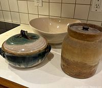 Three ceramic kitchen items: large beige speckled bowl, brown storage jar with lid, and blue-green glazed lidded casserole pot with handles.