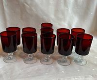 Eleven cranberry glass goblets arranged together showing their rich red color and clear stems against a white backdrop.