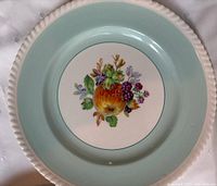 Close-up of aqua/green rimmed dinner plate with painted fruits center. Shows scalloped edge and crazing on glaze.