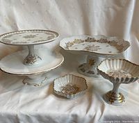 Five pedestal cake plates and dishes arranged on ivory fabric, showing gold floral patterns and gold trim on white porcelain.