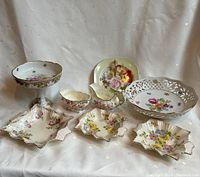 Full view of the lot showing a collection of porcelain and bone china such as lattice bowl, trinket plates, pedestal bowl, creamer, and sugar bowl all decorated with floral patterns and gold trim.