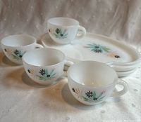 Front angled view of 4 white glass mugs and 4 matching oval plates stacked behind them. All pieces feature a pastel mid-century floral pattern.