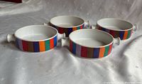 Four white ceramic bowls with bold colorful vertical stripes and knob handles grouped together on a white surface.
