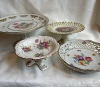 View of four porcelain items: three pedestal cake stands and one serving bowl with floral and gold embellishments on white background.
