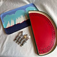 Tray set consisting of a rectangular metal serving tray with a sailboat motif, a wooden tray painted as a watermelon slice, and four cheese spreaders with striped handles.