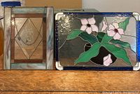 Two stained glass panels displayed on a wooden surface, showing one with pink flowers and green leaves and the other with etched floral motif and hanging chain.