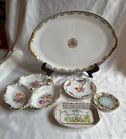 Photo showing five vintage china serving pieces arranged on a white cloth background, including an oval platter, divided dish with handle, two small round plates, and a rectangular dish with painted floral landscape.