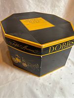 Hexagonal Dobbs Fifth Avenue hat box with yellow trim and horse-drawn carriage illustration on sides.