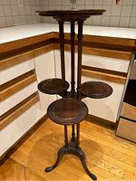 Wood plant stand in corner showing the full height and top surface along with three lower small round shelves on slender vertical supports.