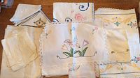 Lot of assorted linens with embroidered floral and geometric designs, white and cream base fabric, lace and scalloped edges, folded and displayed on wooden table.