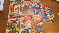 Lot of seven assorted Spiderman universe comic books displayed on a wooden surface, showing colorful covers and various comic book titles.