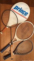 Four badminton rackets lying on wooden floor with one racket inside a Prince JR Mini white cover.