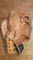 Worn brown leather baseball glove lying on a wood surface showing outer side with laces and wrist strap with label.