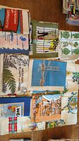 Full overhead view of eight decorative towels laid flat or folded, showing various printed designs including medicinal plants, trees, giraffe, offshore oil rig, city landmarks, and flags.