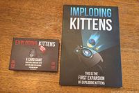 Photo showing both the original Exploding Kittens card game box and the Imploding Kittens expansion box placed side by side on a wooden table.