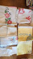 Top view of multiple folded linens with varied embroidery and lace edges, showing floral and geometric patterns in pink, red, yellow, blue, and green threads.