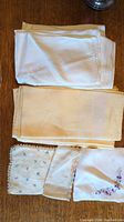 Stack of various folded napkins including plain white and cream colored linens, plus smaller embroidered pieces laid on wooden surface.