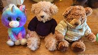 Three stuffed animals on a wooden surface: a rainbow-colored unicorn, a beige teddy bear in purple sweater, and a brown teddy bear in beige speckled sweater