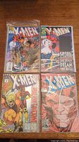 Four X-Men comic books laid out on wooden surface in clear protective sleeves. Covers show various characters and titles.