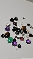 Photo showing multiple polished and faceted mixed natural gemstones scattered on a white background including amethyst, tiger's eye, onyx variants, crystal quartz and more.