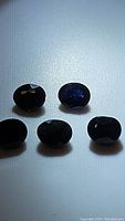 Five oval cut blue sapphire gemstones arranged in two rows on a white background, showing deep blue color and visible inclusions.