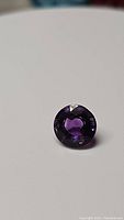 Photo of a single round cut deep purple amethyst gemstone on a plain white background.
