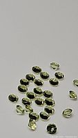 Cluster of small oval-shaped apple green peridot gemstones with reflective surfaces, showing polished and faceted cuts, against a light background.