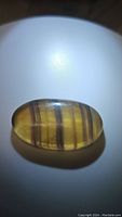 Photo of oval cabochon natural fluorite gemstone with visible yellow and purple banding on a light background.