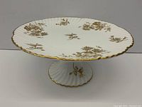 Side angle view of the porcelain cake dish showing scalloped gold edge and pedestal with gold floral decoration.