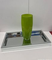 Bright green glass vase standing centered on a rectangular mirrored serving tray with handles, showing full length of both items.