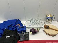 Photo of entire lot showing graduation gowns, CCM garment, tambourine with wooden spoon, Fitz & Floyd eggplant dish, glass containers, glass baking dish, calculators, and a small blue case.