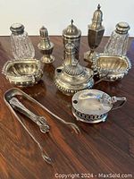 Full grouping on wood surface showing two open salt cellars, two pairs of shakers, three lidded pots and two tongs