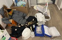 Wide view of the lot showing gray jacket with fur hood, folded baby clothes, carriers, baby bath tub, and blue zippered bag