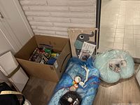 Lot overview showing cardboard box with assorted items, nursing pillow, wrapped items, and toys laid out on floor.