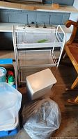 White metal mesh two-tier cart, white plastic garbage can with lid, and coiled rope placed on floor under a shelf
