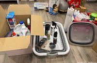 Photo showing box open with two dryer balls in packaging, Clorox bleach bottles, and other liquids inside box, with collapsible basket, shoe shine kit, clothespins, and other cleaning items in background.