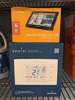 Boxes of Garmin DriveSmart 50 and Emerson Sensi thermostat stacked on a shelf, showing front product packaging with branding and device images.