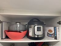 Shelf with Instant Pot Duo 60 V3, clear salad spinner, red and stainless steel bowls, kitchen scale in box, multiple cutting boards and a tray visible.