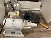 White Champion 2000+ juicer with black cover, covered wok with wooden handle, metal mesh strainer lid, and instruction manuals placed on hardwood floor.