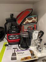 Front view of Ninja Professional Blender 1000 with black and red base and clear pitcher, two stovetop espresso makers, Krups coffee grinder, and two insulated coolers in the background. Ninja blender manuals and power cord also visible.