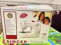 Front of the box showing the Singer Prelude sewing machine and main features like an instructional DVD, easy threading, powerful motor, automatic buttonholes, three presser feet, accessory storage, and free arm.