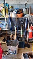 Full view of brooms, shovels, brushes, bucket and pylons arranged together in a storage area.